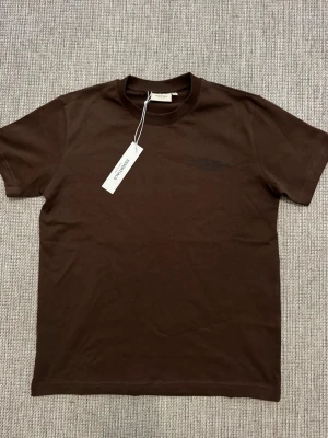 Brun Essentials T-shirt från Fear of God - Snygg brun T-shirt från Fear of God Essentials med tryck på ryggen. Klassisk passform med rund hals och korta ärmar. Tillverkad i mjuk bomull, perfekt för en clean och stilren look.
