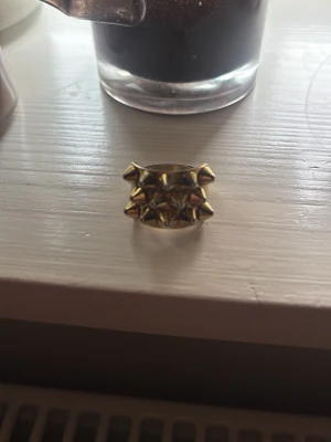 Guldig ring med nitar Edblad  - Cool ring i guldmetall med flera upphöjda nitar i ett rutmönster. Ringen har en bred design och en edgy känsla som sticker ut. Perfekt för dig som gillar statement-smycken och vill addera attityd till din stil.
