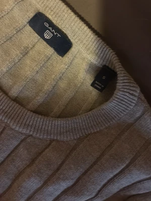 Ljusgrå stickad tröja från GANT - Snygg ljusgrå stickad tröja från GANT med ribbstickat mönster och rund halsringning. Tröjan har en liten broderad logga på bröstet och är tillverkad i bomull. Perfekt för lager-på-lager och enkel att matcha.