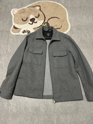 Grå overshirt med fickor från H&M - Stilren grå overshirt från H&M med två bröstfickor och dragkedja framtill. Overshirten har klassisk krage och raka ärmar med knapp vid ärmslut. Materialet är mjukt och känns lite ulligt, perfekt för lager på lager under kyliga dagar.
