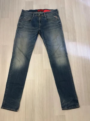 Slim fit blå jeans med slitningar - Snygga slim fit jeans i mellanblå denim med lätt slitna detaljer och klassisk femficksdesign. Jeansen har dragkedjegylf och röda detaljer på insidan av linningen. Perfekta för en avslappnad och trendig look.