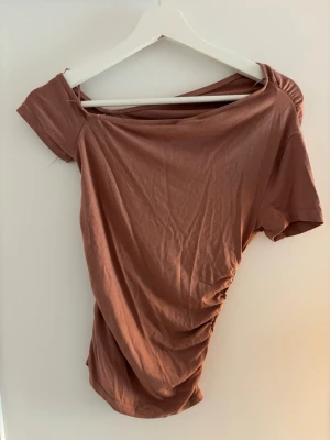 Brun/röd offshoulder topp - Trendig brun topp i mjuk bomull med offshoulder-design och kort ärm. Snygga rynkdetaljer på sidan ger en cool och avslappnad look. Perfekt för dig som gillar en lite annorlunda och modern stil. Nästan aldrig använd 