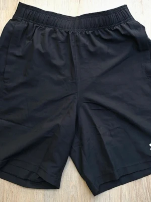 Svarta träningsshorts från Under Armour - Svarta träningsshorts från Under Armour med elastisk midja och logga på benet. Lätta och bekväma, perfekta för gymmet eller löpning. Klassisk design med diskret logotyp och bra passform.