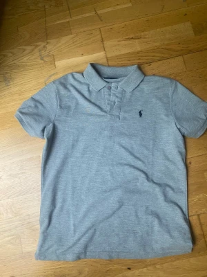 Grå pikétröja från Ralph Lauren - Klassisk grå pikétröja från Ralph Lauren med korta ärmar och krage. Tröjan har knappar framtill och den ikoniska broderade loggan på bröstet. Perfekt för en stilren och avslappnad look.