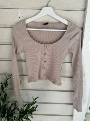 Beige ribbad topp från Gina Tricot storlek: S - Snygg beige ribbad topp från Gina Tricot med lång ärm och croppad passform. Toppen har rund halsringning och dekorativa knappar framtill. Perfekt att styla med jeans eller kjol för en trendig look.