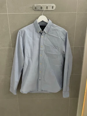 Ljusblå skjorta från Bondelid - Snygg ljusblå skjorta från Bondelid med klassisk button-down krage och bröstficka. Skjortan har långa ärmar och vita knappar framtill. Perfekt för en stilren och avslappnad look. Jävligt dåliga bilder! Orkar inte ta bättre. Kan skrivas ifall bra bilder krävs.