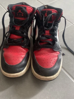 Air Jordan 1 Mid röd svart vit - Säljer ett par Air Jordan 1 Mid sneakers i klassisk röd, svart och vit färg. Skorna har högt skaft, svart snörning och ikoniska Jordan-loggor på plös och sida. Ovandelen är i läder och designen är riktigt tidlös för dig som gillar streetwear.
