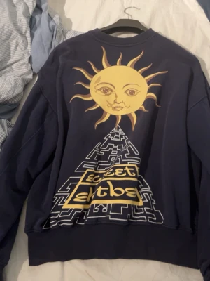 Mörkblå sweatshirt sweet skeatboards - Cool mörkblå sweatshirt från sweet skeatboards med tryck på bröstet och stor grafisk design på ryggen med sol och pyramid i gult och vitt. Tröjan har rund hals, ribbade muddar och är tillverkad i mjukt bomullsmaterial. Perfekt för en avslappnad streetwear-look.