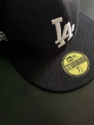  New Era keps - Svart keps från New Era 59FIFTY med LA Dodgers-logga framtill i vitt och broderad 1988 World Series-detalj på sidan. Klassisk flat peak och strukturerad passform. Materialet är bomullstwill och kepsen har en guldfärgad etikett på skärmen.