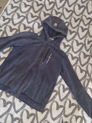 Hoodie från Sail Racing unisex - Mörkblå hoodie från Sail Racing med dragkedja framtill och en snedställd bröstficka med logga. Luva med snören. Mjuk och skön kvalitet, perfekt för chill dagar. Ljusa fläckar finns- ser ut som blekt. I övrigt fint skick