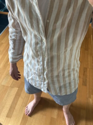 Beige och vit randig linneskjorta - Snygg skjorta i beige och vitt med vertikala ränder. Skjortan har klassisk krage, knäppning framtill och långa ärmar. Tillverkad i luftigt linne, perfekt för varmare dagar. Enkel och stilren design som passar till många outfits.