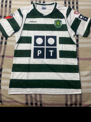 Sporting Lissabon fotbollströja Ronaldo - Grönvit randig fotbollströja från Reebok med Sporting Lissabon-logga och sponsortryck. På ryggen står det C. Ronaldo och nummer 28. Tröjan har korta ärmar och klassisk v-ringning. Perfekt för dig som älskar fotboll och vill ha en ikonisk supportertröja. Priset är ej hugget i sten!