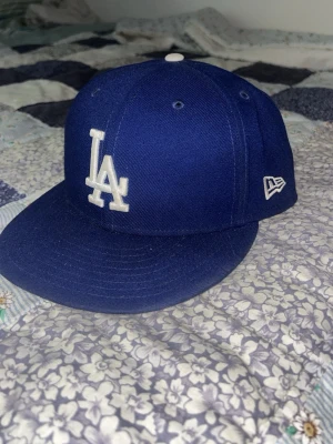 Blå LA Dodgers keps från New Era - Snygg blå keps från New Era med LA Dodgers-logga broderad i vitt framtill. Klassisk flat brim och broderad MLB-logga baktill. Perfekt för dig som gillar sportig streetstyle och vill ha en ikonisk look.