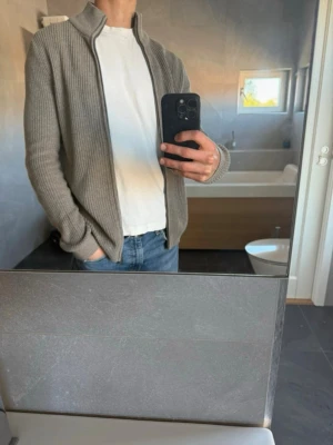 Beige ribbstickad ziptröja - Snygg beige ribbstickad ziptröja med hög krage och dragkedja. Tröjan har en avslappnad passform och är perfekt att slänga över en t-shirt. Tillverkad i mjukt material som känns skönt mot huden. Använd ca 5 gånger.