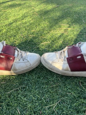 Valentino vita sneakers med vinröda detaljer - Snygga vita sneakers från Valentino med breda vinröda läderdetaljer över snörningen. Klassisk låg modell med rund tå och vita skosnören. Perfekta för dig som vill ha en stilren men ändå unik look.