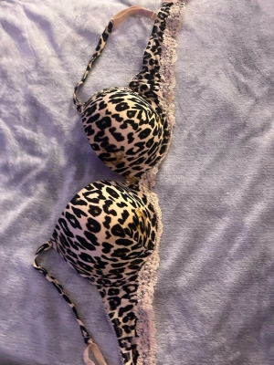 Leopardmönstrad push-up bh med spets - Snygg push-up bh med leopardmönster i svart och beige. Bh:n har vadderade kupor och dekorativ vit spets längs nederkanten. Justerbara axelband för perfekt passform. Perfekt för dig som gillar djurmönster och vill ha något extra i underklädeslådan.