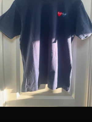 Mörkblå Comme des Garçons t-shirt - Säljer en mörkblå t-shirt från Comme des Garçons med två hjärtan i rött och blått broderade på bröstet. Klassisk passform och korta ärmar. Tillverkad i mjuk bomull, perfekt för en clean och stilren look.