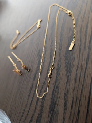 YSL-inspirerat smyckeset i guld - Säljer ett snyggt smyckeset i guldfärgad metall med YSL-inspirerade detaljer. Setet innehåller ett halsband, ett armband och ett par örhängen, alla med tunna kedjor och YSL-logga som hänge. Perfekt för dig som gillar trendiga accessoarer med lyxig känsla.
