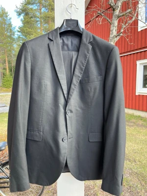 Svart kostym från Jack & Jones - Studentkostym! Stilren svart kostym från Jack & Jones Premium med klassisk passform och två knappar framtill. Kavajen har tre fickor på framsidan och är helfodrad. Perfekt för en dressad look och passar till många olika tillfällen.   Storlek 46  Midjemått: 83cm Beninnerlängd: 77cm  Rygglängd från nacke och neråt: 73cm Bredd över ryggen, armhåla till armhåla: 50cm  Bra skick. Rök- och djur-fritt hem.