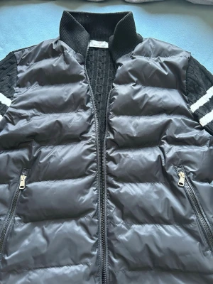 Svart pufferjacka från Moncler - Snygg svart pufferjacka från Moncler med stickade ärmar och vita ränder. Jackan har dragkedja framtill, två fickor med dragkedja och ribbad krage. Perfekt för dig som vill ha en sportig och trendig look.