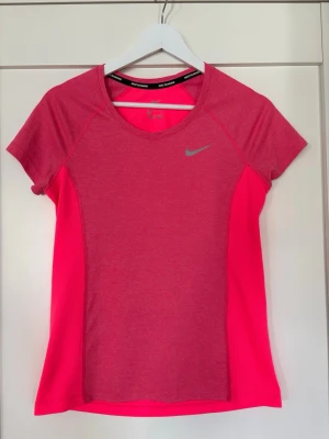 Nike Dri-Fit rosa tränings-tshirt - Nike Dri-Fit tränings-tshirt i starkt rosa med meshpaneler för extra ventilation. T-shirten har korta ärmar, rund hals och reflekterande Nike-logga på bröstet. Storlek S