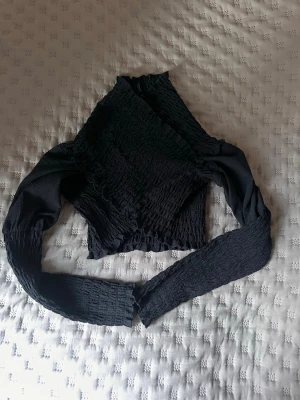 Svart croppad topp från NA-KD - Trendig svart croppad topp från NA-KD med långärmad design och smockad struktur. Toppen har en åtsittande passform och är perfekt för att skapa en cool och stilren look. Materialet är mjukt och stretchigt för extra komfort.