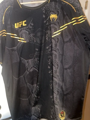 Svart UFC t-shirt från Venum - Säljer en svart UFC t-shirt från Venum med coolt ormmönster och gula detaljer. Tröjan har tryckta loggor på bröstet och axlarna samt sportig passform. Perfekt för träning eller att representera din favoritorganisation.