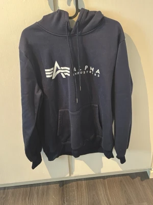 Mörkblå hoodie från Alpha Industries - Mörkblå hoodie från Alpha Industries med vit logga och tryck framtill. Klassisk känguruficka och justerbar dragsko i huvan. Mjuk bomullsblandning och avslappnad passform, perfekt för en chill och stilren look.