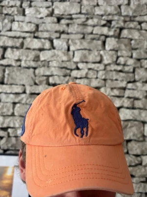 Orange keps från Polo Ralph Lauren - Säljer en snygg orange keps från Polo Ralph Lauren med klassisk blå broderad logga framtill och en stor blå siffra 3 på sidan. Kepsen har böjd skärm och justerbar rem bak. Perfekt för dig som vill ha en sportig och trendig look.