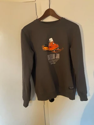Mörkgrå sweatshirt Ockelbo 300 - Mörkgrå sweatshirt med tryck av en orange Ockelbo 300 snöskoter och text på bröstet. Rund halsringning och långa ärmar. Oanvänd