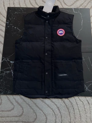 Svart dunväst från Canada Goose - Stilren svart dunväst från Canada Goose med klassisk logotyp på bröstet och två stora fickor framtill. Västen har hög krage, dragkedja och knappar samt diskret Canada Goose-märke på ena fickan. Perfekt för lager-på-lager och streetstyle.kommer med Canada goose påse också 