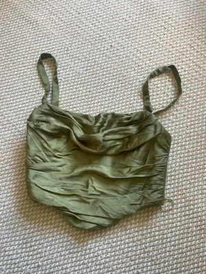 Olivgrön korsett-topp med drapering - Snygg olivgrön korsett-topp med draperad framsida och smala axelband. Toppen har en figurnära passform och är tillverkad i ett glansigt material som ger en lyxig känsla. Perfekt att styla med jeans eller kjol för en trendig look. 