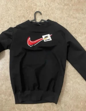 Svart sweatshirt från Nike - Svart sweatshirt från Nike med röd och vit swoosh-logga på bröstet och en liten patch med texten 'Nike Sportswear'. Klassisk rund halsringning och ribbade muddar vid ärmslut och nederkant. Perfekt för en sportig och avslappnad stil.
