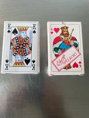 Två kortlekar i originalförpackning - Säljer två klassiska kortlekar i originalförpackning. Ena asken har motiv av spader kung i svart, vitt och gult, den andra har hjärter kung med färgglad design och texten 'Svenska Spelkort'. Perfekt för kortspel med vänner.