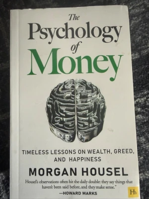 The Psychology of Money bok - Säljer boken 'The Psychology of Money' av Morgan Housel. En pocketbok med vit framsida, gröna och svarta bokstäver samt illustration av en hjärna gjord av sedlar. Boken handlar om pengar, beteende och lycka.