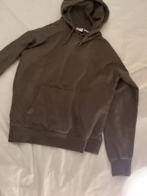 Olivgrön hoodie från Gina Tricot - Snygg olivgrön hoodie från Gina Tricot med klassisk känguruficka och dragsko i huvan. Mjuk insida och ribbade muddar vid ärmslut och nederkant. Perfekt för en avslappnad och trendig stil.