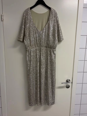 Glittrig silverklänning med v-ringning - Snygg glittrig klänning i silver med v-ringning och korta ärmar. Klänningen har paljetter över hela tyget och en rak, något lös passform som ger en cool och festlig look. Perfekt för dig som vill sticka ut med stil. Upplever den som lite större i storleken 