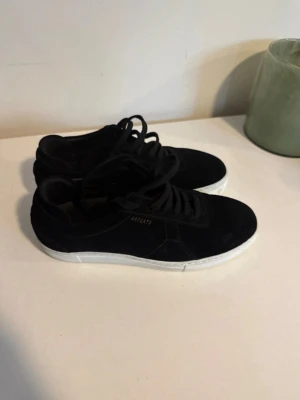 Nya svarta sneakers från Axel Arigato - Snygga svarta sneakers från Axel Arigato med vit sula och diskret logga på sidan. Använda en gång! Skorna har klassisk låg modell, svarta skosnören och är tillverkade i mocka. Perfekta för en stilren och trendig look.