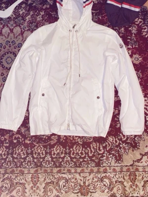 Vit vindjacka från Moncler - Snygg vit vindjacka från Moncler med huva och dragsko. Jackan har dragkedja framtill, två fickor med tryckknappar och Moncler-logga på ärmen. Materialet är lätt och vattenavvisande, perfekt för blåsiga dagar. Röd och blå randdetalj på huvan ger en sportig touch.