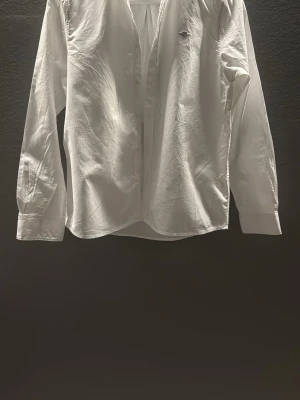 Vit skjorta från GANT - Klassisk vit skjorta från GANT med knappar framtill och lång ärm. Skjortan har en stilren design med liten logga på bröstet och är tillverkad i mjuk bomull. Perfekt för en clean och tidlös look.