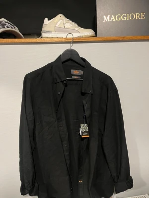 Svart linneskjorta från Morris - Säljer en svart linneskjorta från Morris med klassisk passform och button down-krage. Skjortan har långa ärmar och diskreta knappar framtill. Perfekt för dig som gillar stilrena och tidlösa plagg i ett luftigt material.