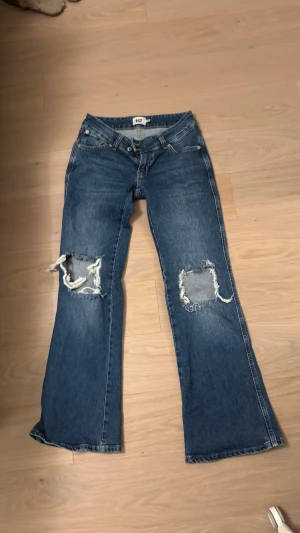 Blå slitna bootcut jeans  - Säljer ett par blå bootcut jeans från lager 157 med stora slitningar över båda knäna. Jeansen har klassisk femficksmodell, låg midja och råa kanter vid bensluten. Perfekta för en avslappnad och trendig look.