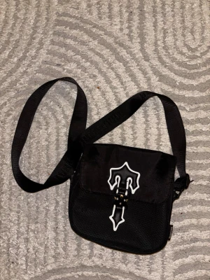 Svart axelväska med vit detalj - Snygg svart axelväska med justerbar rem och unik vit grafisk detalj på locket. Väskan har en fyrkantig form och stängs med spänne framtill. Perfekt för dig som vill ha en stilren men ändå cool accessoar.
