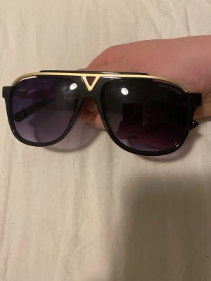Svarta Louis Vuitton solglasögon - Snygga svarta solglasögon från Louis Vuitton med guldfärgade detaljer och mörka glas. Klassisk pilotformad båge som ger en lyxig och trendig vibe. Perfekta för att lyfta vilken outfit som helst.