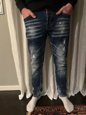 Dsquared2 blå skinny jeans - Säljer ett par blå skinny jeans från Dsquared2 med slitningar, vita och röda färgstänk samt stora patchdetaljer bak. Jeansen har en tvättad look och smal passform, perfekta för dig som gillar streetwear och vill sticka ut.