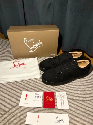 Svarta sneakers från Christian Louboutin - Snygga svarta sneakers från Christian Louboutin med klassisk röd sula och diskret logga. Skorna har en stilren design i mocka och läder, svarta skosnören och lågt skaft. Perfekta för dig som vill ha en exklusiv och trendig look.