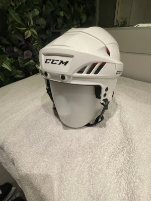 Vit hockeyhjälm från CCM stl. Medium  - Säljer en vit hockeyhjälm från CCM med svart hakrem och ventilationshål. Hjälmen har en modern design med spännen. Nytt skick då den knappast har blivit använd.