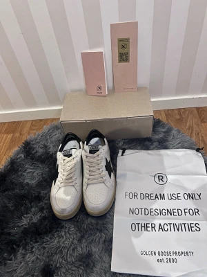 Golden Goose skor  - Snygga vita sneakers från Golden Goose med svart stjärndetalj på sidan och svart häl. Skorna har GGDB-logga på plösen, perforerad tå och beige sula. Tillverkade i läder och har klassisk snörning. Kommer med originalpåse och låda.