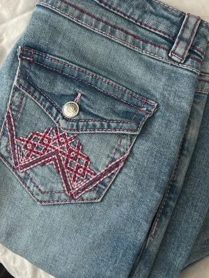 Blå jeans med rosa sömmar & brodyr - Säljer ett par ljusblå jeans med rosa kontrastsömmar och snygg broderad detalj på bakfickan. Jeansen har slitningar på ena benet för en trendig look och klassisk femficksmodell. Perfekta för dig som gillar unika detaljer och streetstyle.