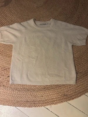 Beige stickad t-shirt från lager 157 - Kortärmad stickad t-shirt i beige från lager 157. Tröjan har rund halsringning och ribbade kanter vid ärmslut och nederkant. Perfekt för en avslappnad och stilren look. Materialet ser mjukt och bekvämt ut.